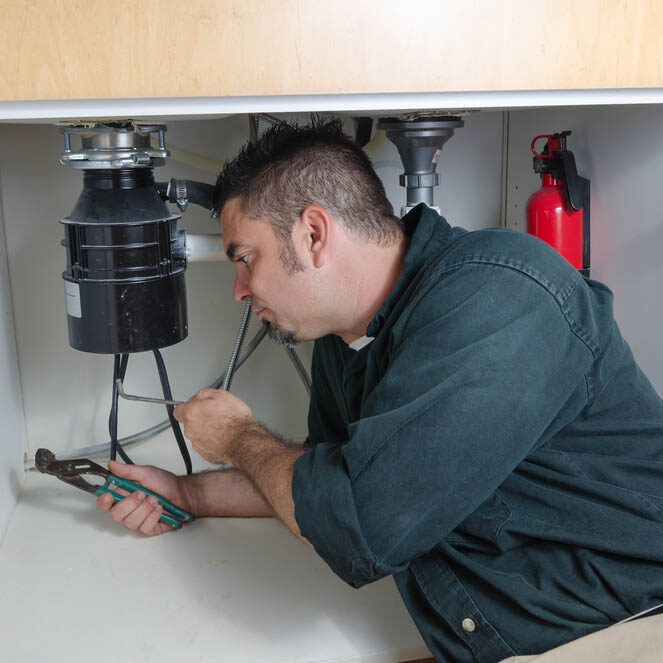 A plumber laying under a house hold sink working on a garbage disposal.