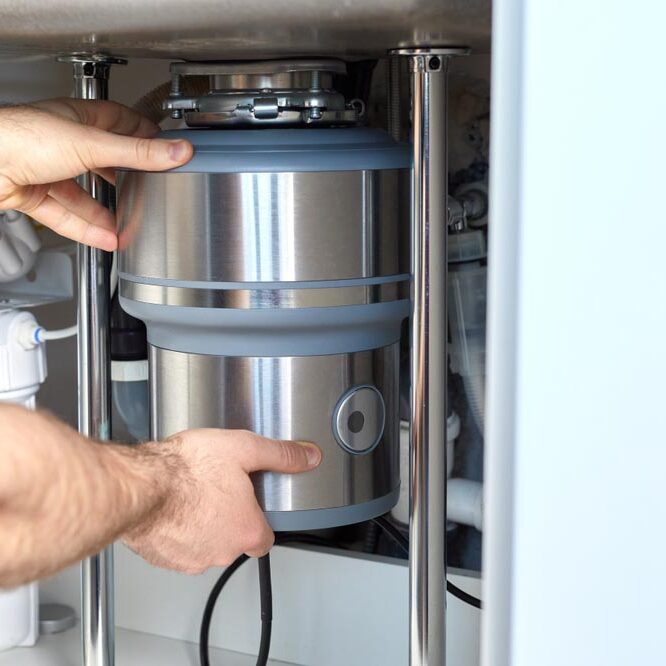 A plumber with gloved hands installs a household waste shredder for the kitchen sink. A technician installs or repairs a household waste shredder in the kitchen.